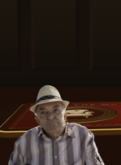 An elderly man in a white hat and striped shirt sits in a dimly lit room in front of a glowing table with a game-like board.