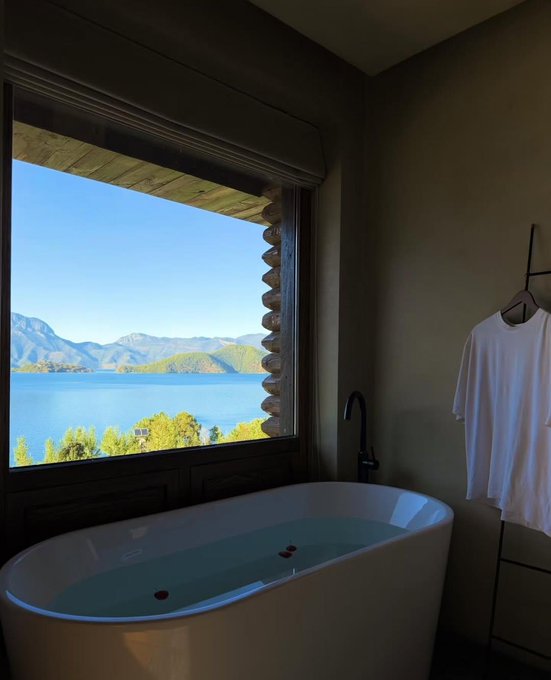 A bathroom interior featuring a white bathtub filled with water, a white t-shirt hanging on a rack, and a window view of mountains and a lake.