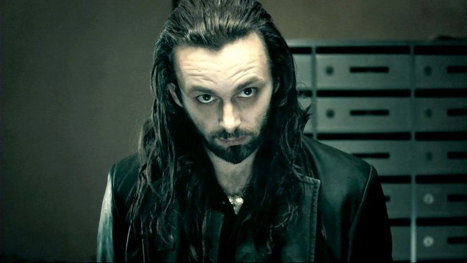 A cinematic portrait shows a man with long dark hair and a goatee, wearing a black leather jacket, staring intensely at the camera.
