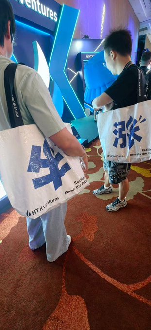 Two attendees at a lively HTX Ventures booth carry white tote bags featuring bold blue Chinese characters and the slogan "Revive the bull," with the hashtag #HTXventures printed below. The "Ventures" signage and "Just Buy" branding on display walls confirm this is an HTX promotional event, likely TOKEN2049 as referenced in the caption, where guests can snag the creatively designed merch early before supplies run out.