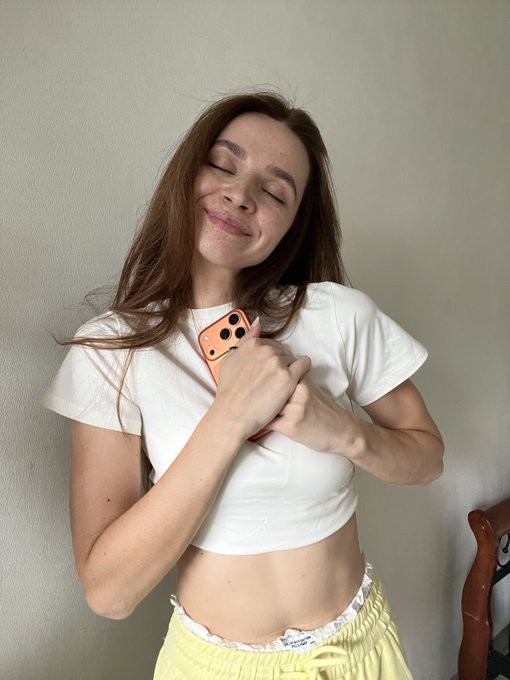 A smiling woman with closed eyes hugs an orange iPhone to her chest, wearing a white t-shirt and yellow pants.