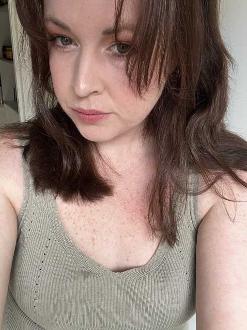 A selfie of a young woman with shoulder-length wavy brown hair, freckles across her chest and face, wearing a sage green ribbed tank top.