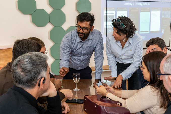 The image shows a collaborative workshop scene where participants gather around a table with smartphones, while a screen in the background displays "Activity 3 - Verify Beta-iD." Two facilitators, one in a blue shirt and another in a white shirt, engage closely with the group, illustrating a hands-on session exploring blockchain-based identity verification solutions for public institutions.