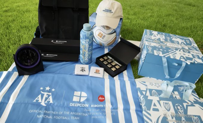 A collection of official merchandise and accessories for the Argentina National Football Team laid out on grass, including a cap, scarf, a Deecoin branded water bottle, and boxes, with a label reading 'OFFICIAL PARTNER OF THE ARGENTINA NATIONAL FOOTBALL TEAM 2026.07.15'.