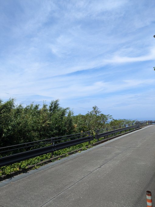 A scenic coastal road stretches into the distance, bordered by lush green vegetation and a metal guardrail, while a stunning blue sky with wispy clouds fills the upper portion of the frame. The image captures the serene and expansive view from the JPYC Aogashima office, showcasing the island's natural beauty and picturesque surroundings.