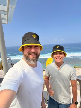 A man and a boy take a selfie by the ocean, both wearing matching black and yellow bucket hats and white shirts.
