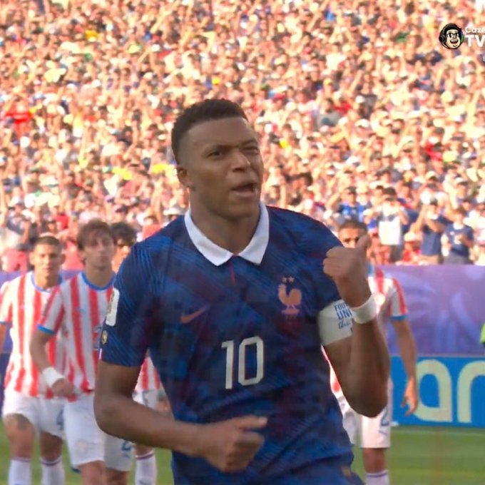 A French soccer player wearing jersey number 10 and a captain's armband marked "UNI" celebrates a goal with a determined expression and thumbs-up gesture, while opposing players in red and white striped uniforms stand in the background amid a packed stadium. The image appears to show Kylian Mbappé during a match between France and Paraguay, fitting the post's reference to a new anthem moment in football.