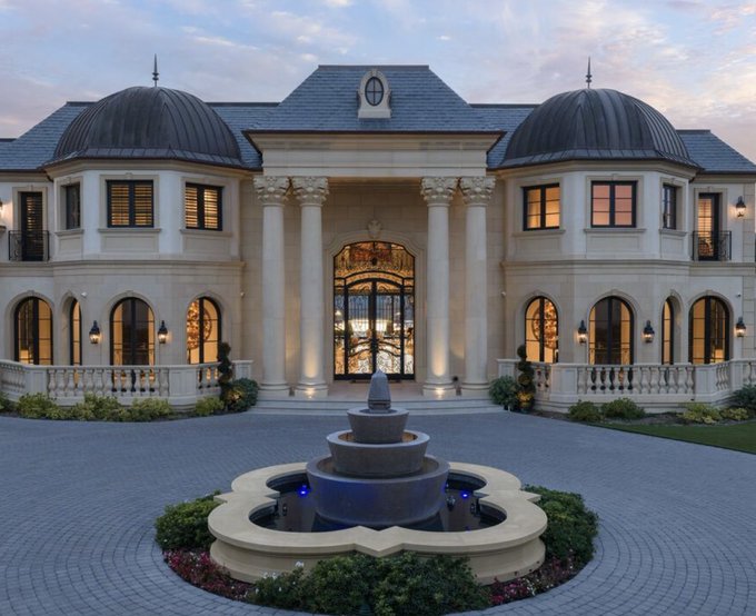 A grand, palatial mansion featuring classical architecture with columns, domed roofs, arched windows, and a tiered fountain in the circular driveway.