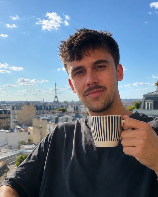 A young man with a mustache sips from a striped mug while posing for a photo on a rooftop, with the Eiffel Tower visible in the Parisian skyline behind him.
