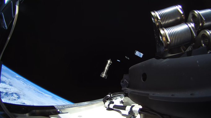 A photograph taken from space showing a satellite being deployed, with Earth visible in the lower left and the black void of space surrounding mechanical equipment and a small free-floating object.