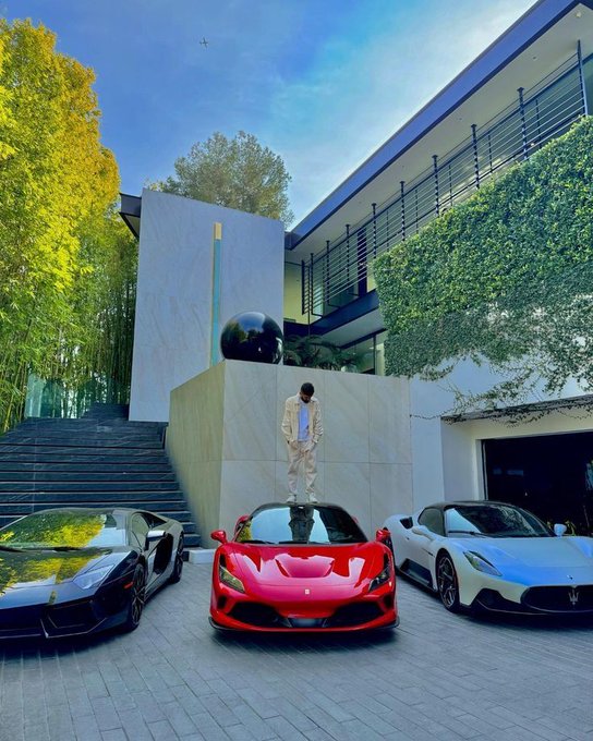 A man in a tan suit stands on top of a red Ferrari sports car parked between a black Lamborghini and a white Maserati in front of a modern luxury home.