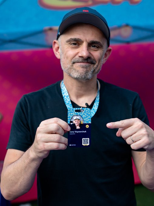 A smiling middle-aged man wearing a black cap and t-shirt holds up a blue lanyard with a credential card that reads 'Dan Vaynchek' and displays a QR code.
