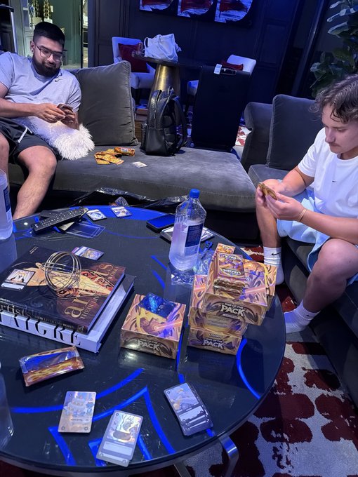 Two men relaxing on a grey couch in a lounge area while playing with Pokémon trading cards, with several booster boxes and packs of cards scattered on a round black table.