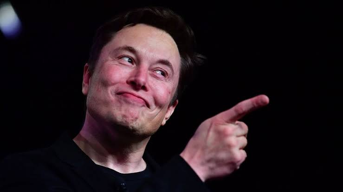 A close-up photo of Elon Musk smiling and looking to the side while pointing his finger off-camera against a dark background.