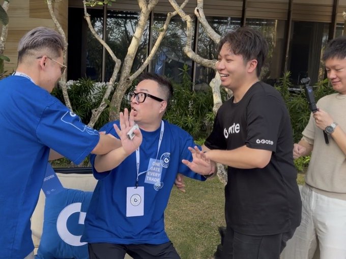 Four people at a Gate event pose dramatically, with one man in a blue shirt clutching his chest in mock distress while two others reach toward him and a fourth holds a microphone. The man being "ambushed" wears a blue t-shirt with "GATE IS EVERYTHING" text and a badge featuring the letter "C," while the others wear black Gate-branded shirts, humorously illustrating the post's claim that 倪大 (Ni Da) was ambushed by 林克, Unicorn, and Ric at the Gate event.