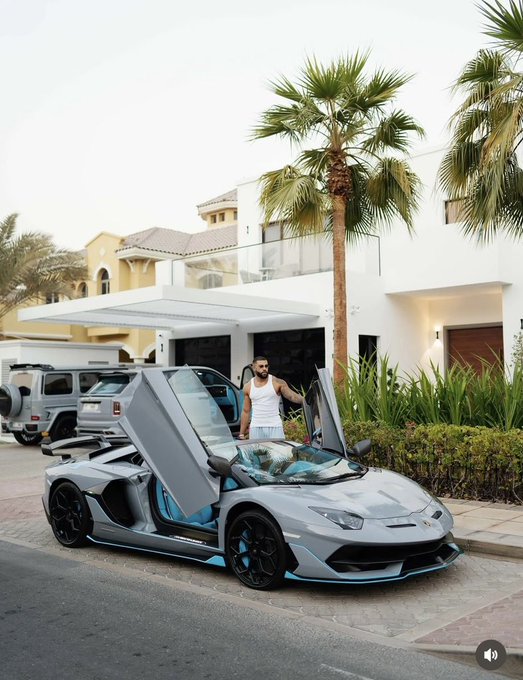 The image features a man standing next to a luxury sports car, which appears to be a Lamborghini Huracan. The car has its doors open, revealing its interior. In the background, there are two other cars parked nearby - one white and another black. A palm tree can also be seen in the scene. The post seems to be discussing the importance of projects that create noise or generate buzz, as well as the significance of quietly building systems and communities for long-term success.