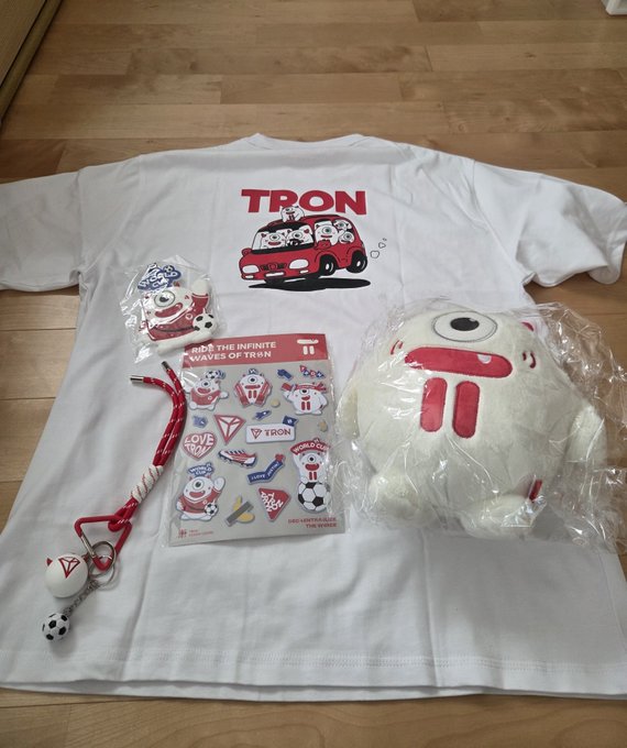 A white T-shirt featuring the "TRON" logo and a red van graphic is laid out on a wooden floor, accompanied by Tron-branded merchandise including a plush toy, keychain, and a sticker sheet labeled "INTO THE INFINITE WAVE OF TRON." The collection showcases the fan's excitement for receiving official TRON DAO merchandise.