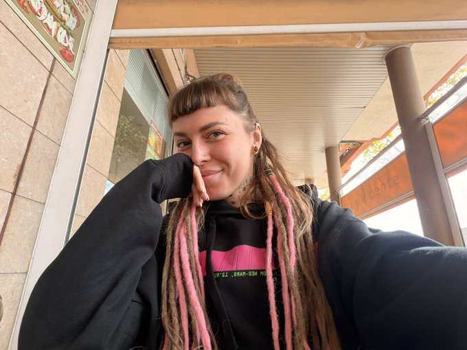 A selfie of a young woman with long dreadlocks featuring pink streaks, wearing a black hoodie, sitting on an outdoor patio with her hand near her face.
