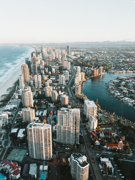 An aerial photograph captures the Gold Coast skyline with high-rise buildings lining the beachfront and the Nerang River, showcasing the coastal city at golden hour.