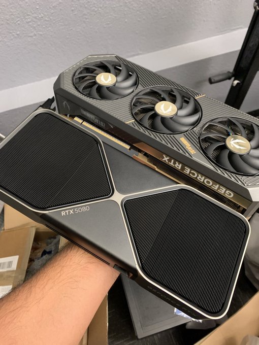 A person's arm holds two high-end graphics cards, with a ZOTAC GeForce RTX 5090 visible on top and another RTX 5080 graphics card below it.
