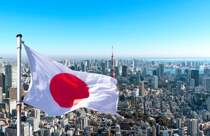 A Japanese flag waves in the foreground against the backdrop of Tokyo's skyline, featuring landmarks like Tokyo Tower, symbolizing Japan's role in pioneering blockchain-based 24/7 instant settlement for stocks and government bonds—another milestone toward fully onchain, always-on financial markets.