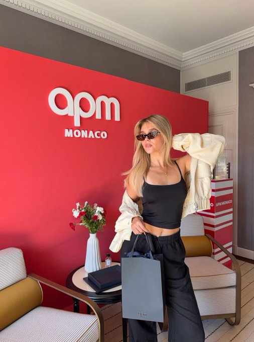 A blonde woman in sunglasses, a black crop top, and an oversized white jacket poses with a shopping bag in front of an 'apm MONACO' red backdrop.