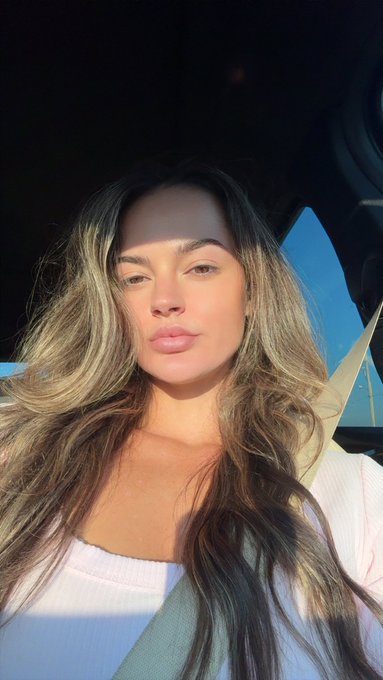 A woman with long wavy brown hair takes a sunlit selfie in a car.