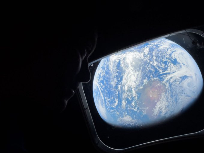 A silhouetted figure gazes through a spacecraft window at the glowing blue planet Earth suspended in the black void of space, embodying the awe-inspiring experience of having "the best window seat in the universe."