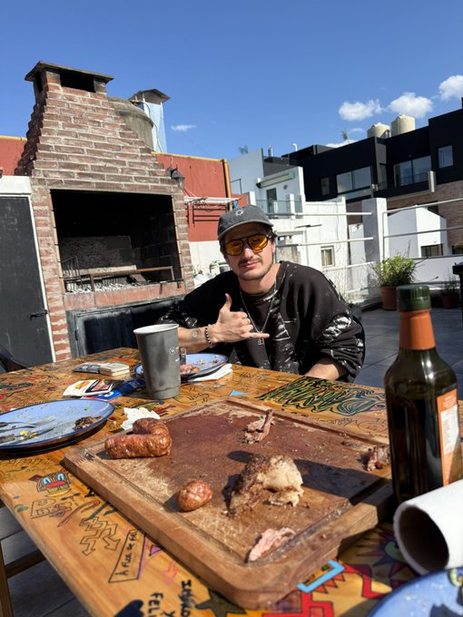 A man in a black shirt, cap, and sunglasses sits at an outdoor table with a wooden cutting board of meats, plates, a cup, and a wine bottle, with a brick grill behind him.