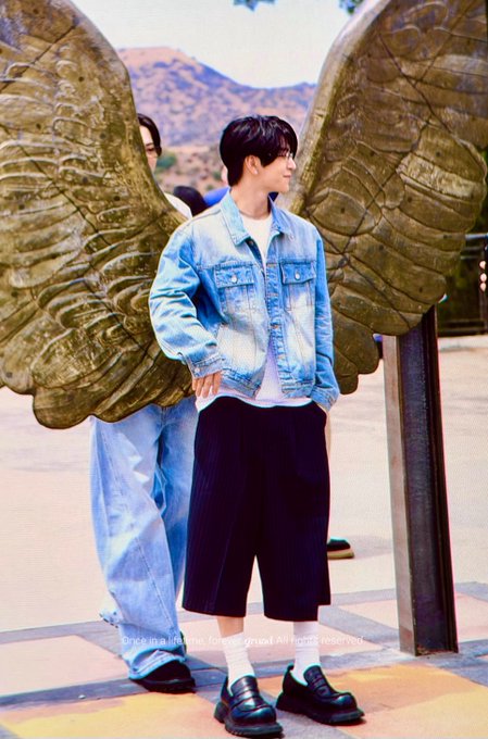 A young man wearing a light blue denim jacket, black shorts, white socks, and black loafers poses in front of large golden wing sculptures, with a caption reading "once in a lifetime" visible at the bottom of the image. The accompanying post hashtags reference the artist Xinlong (鑫隆) and the song/album "One day I'll be famous," connecting the stylish, photogenic shot to his rising celebrity persona and ALD1 (Alphadriveone) project branding.