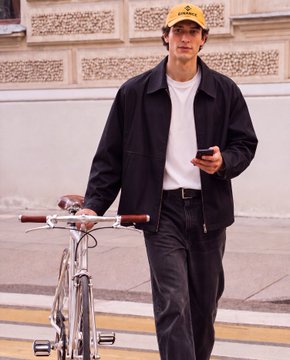 A man wearing a yellow cap, black jacket, white t-shirt, and dark trousers walks alongside his bicycle while looking at his phone, embodying a casual, urban "fit check" aesthetic.