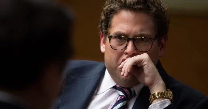 A man with curly brown hair and tortoiseshell glasses, wearing a dark suit, white shirt, and patterned tie, sits with his hand thoughtfully resting against his mouth in what appears to be a formal meeting or hearing setting. His contemplative expression and focused gaze suggest he is intently listening or considering the question posed about Garrett's ownership stake in ZEC.