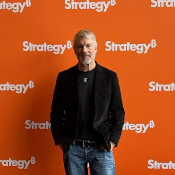 A man with grey hair and a beard stands smiling in front of an orange backdrop featuring repeated white "Strategy" logos.