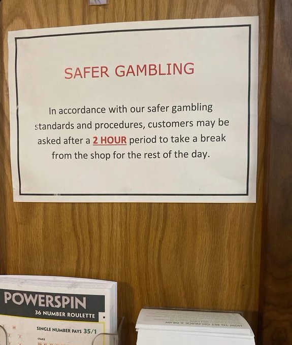 The image shows a wooden door with a sign posted on it that reads "Safer Gambling". Below the sign, there are two books placed next to each other - one titled "Power Spin" and another called "Hyperliquid". The text on the sign is in red and black colors, which stands out against the white background.