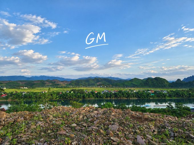 A scenic landscape photograph showing mountains, a river, and a village under a blue sky with clouds, with "GM" (Good Morning) text handwritten in the sky.