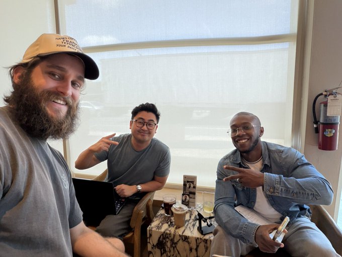 The image features three men sitting around a table at a coffee shop or diner. One of them has a beard and is wearing a hat with "Lloyd's" written on it, indicating that they might be discussing Dogecoin. The other two men are smiling and appear to be enjoying their time together. In the background, there is a window overlooking an outdoor area where a car can be seen parked.