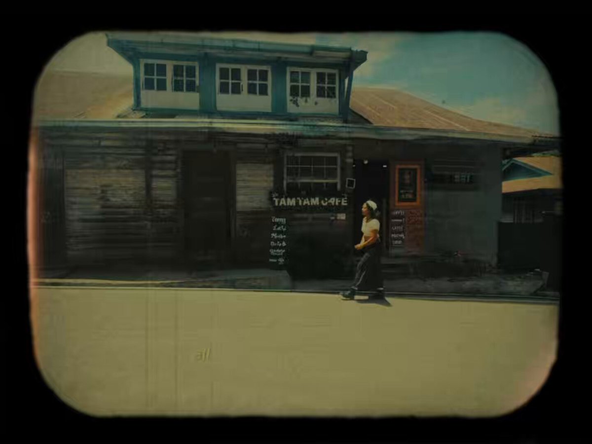 The image features a man walking past an old building with a sign that reads "TAMAM CAFE". The photo has been edited or filtered to give it a vintage appearance, giving it a nostalgic feel. The caption of the image includes some text about a trip down memory lane and mentions that the photo was taken by @Sunrisedefi.