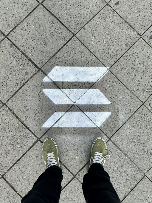 A person's feet are captured in a picture with a line of white squares beneath them that reads "SOLANA!". The individual is wearing green sneakers and black pants while standing on concrete stairs or an asphalt road, possibly outdoors. The presence of this text on the photograph and the person's outfit suggest it may be related to an event or gathering involving cryptocurrency, such as the Solana Summit that has gained attention in recent years due to its potential for disrupting traditional financial systems with innovative technologies like blockchain.
