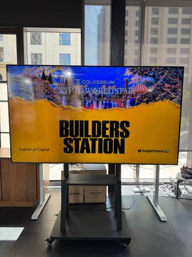 A flat-screen TV on a rolling stand displaying a Crypto World Fair promotional slide with 'BUILDERS STATION' in large text, alongside 'ZothFi at CapMar' and 'Hyperbeam' logos.