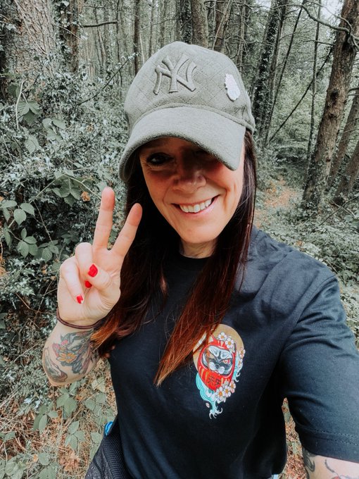 A woman wearing a New York Yankees cap and a black t-shirt with a tiger design, smiling and holding up a peace sign in a forest setting.