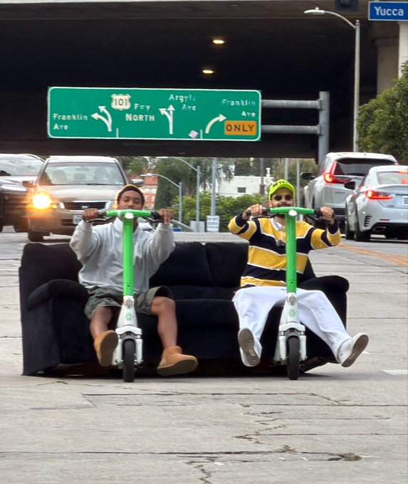 The image shows two people riding a green scooter with a black seat, which appears to be an electric one-seater. They are sitting on top of a couch that has been repurposed as their mode of transportation. The scene takes place in front of a highway sign indicating the direction to Franklin Drive and Franklin Avenue.