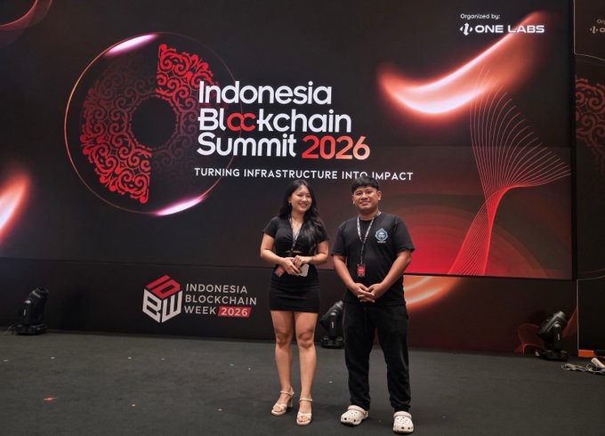 Two people stand smiling in front of a large screen displaying 'Indonesia Blockchain Summit 2026' with the tagline 'TURNING INFRASTRUCTURE INTO IMPACT' at an event organized by ONE LABS.