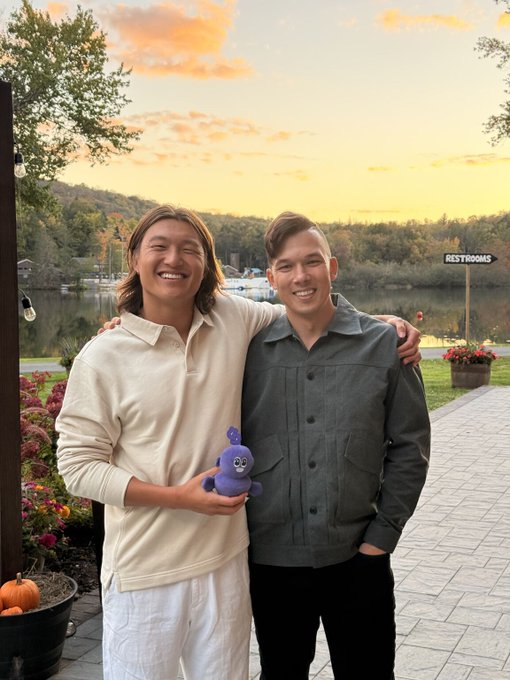 Two men smiling together outdoors at sunset by a lake, with the man on the left holding a purple plush toy.