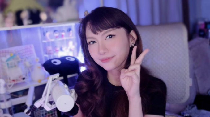 Portrait of a young woman with long brown hair smiling and making a peace sign, sitting in front of a white microphone in a room decorated with collectibles and display cases.