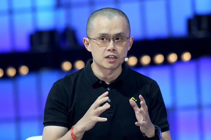 Changpeng "CZ" Zhao, founder of Binance, speaks at a conference while gesturing with his hands, providing visual context for his bullish statement that owning 0.1 Bitcoin will eventually be worth more than a house. He wears a black polo shirt, glasses, and a wristband, with a headset microphone visible.