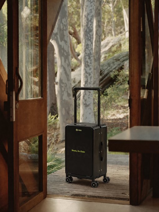 A black carry-on suitcase by NEX with the text 'Shells. On. Chain.' printed on it, positioned in a doorway looking out into a forest with large trees.