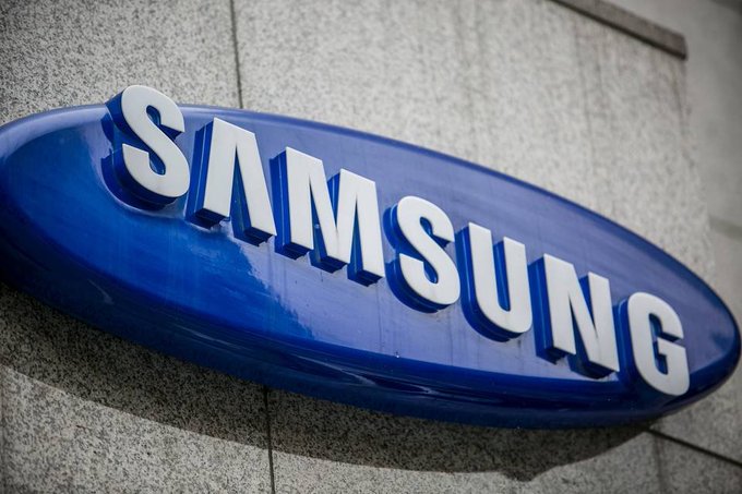 A close-up of Samsung's blue oval logo mounted on a stone building facade, representing the company that plans to integrate native stablecoin features into its 800 million Galaxy smartphones through Samsung Wallet. The bold white "SAMSUNG" lettering on the glossy blue emblem visually anchors the news of the tech giant's move into digital currency services.