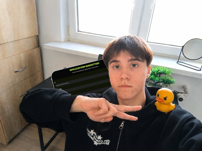 A young man in a black hoodie poses with a peace sign next to a rubber duck perched on his shoulder, with a laptop behind him displaying "APPLICATION SUBMITTED!" — capturing the moment he applied to the @duel_duck ambassador program mentioned in his post.