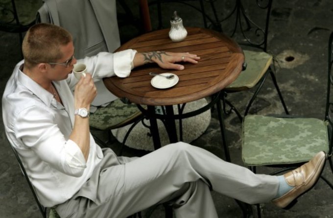 A man in a white shirt and light-colored pants sits relaxed at a small outdoor café table, sipping coffee with his legs casually crossed and stretched out. The image visually embodies the post's warning about market complacency, depicting the kind of calm, unbothered posture investors tend to have right before a sudden and dramatic pullback in altcoins.