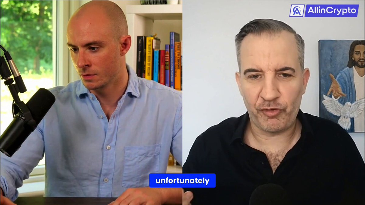 The image shows two men sitting at a desk, with one man wearing a blue shirt and the other wearing black. They are both looking at a computer screen that displays an interview between them. The man in the blue shirt has a microphone in front of him, while the man in the black shirt is seated behind it. Both men appear to be engaged in conversation or discussing something on the computer screen.